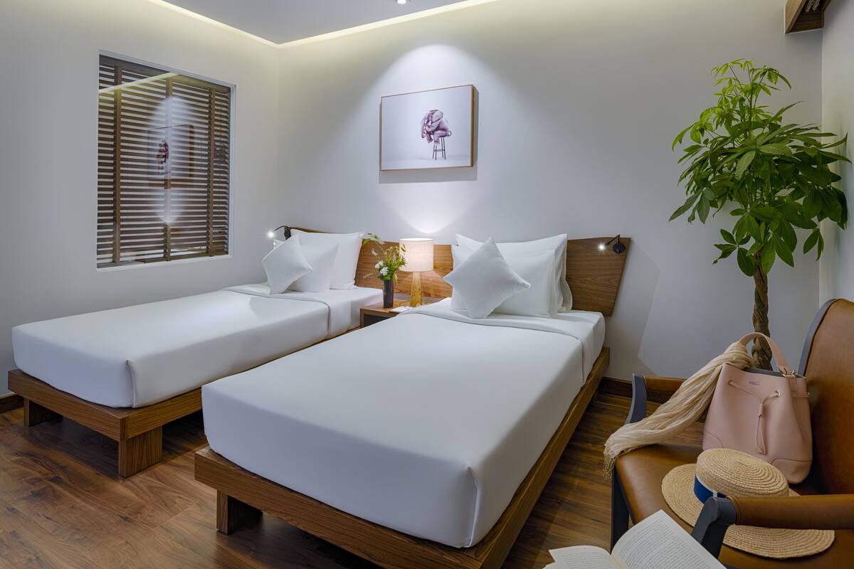 Een hotelkamer met twee enkele bedden, een houten vloer, een nachtkastje met een lamp, een lijst met een afbeelding aan de muur en een potplant.