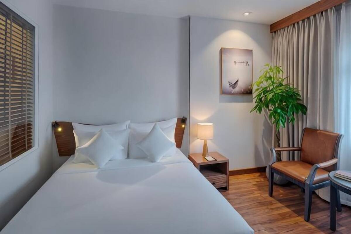 Een hotelkamer met een bed, nachtkastje, lamp, stoel en een grote potplant.