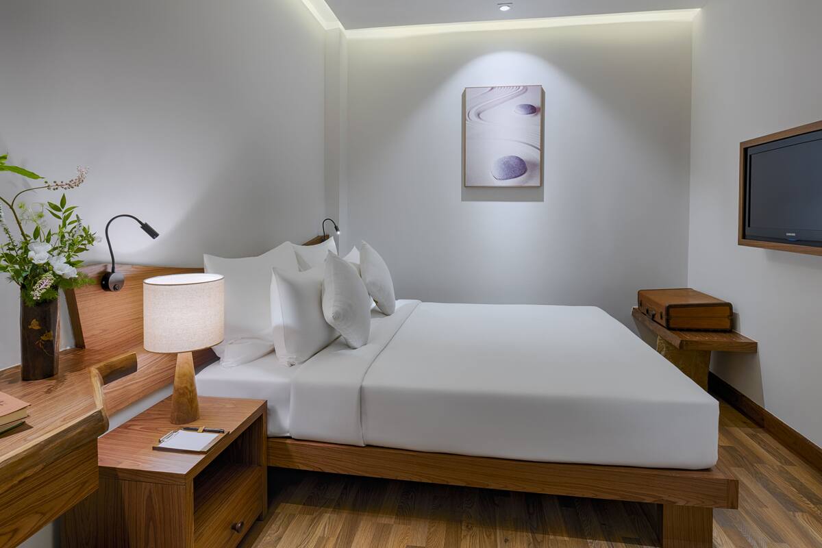 Een moderne hotelkamer met een houten bed, witte beddengoed, een bureau met lamp, een klein nachtkastje en een televisie aan de wand.