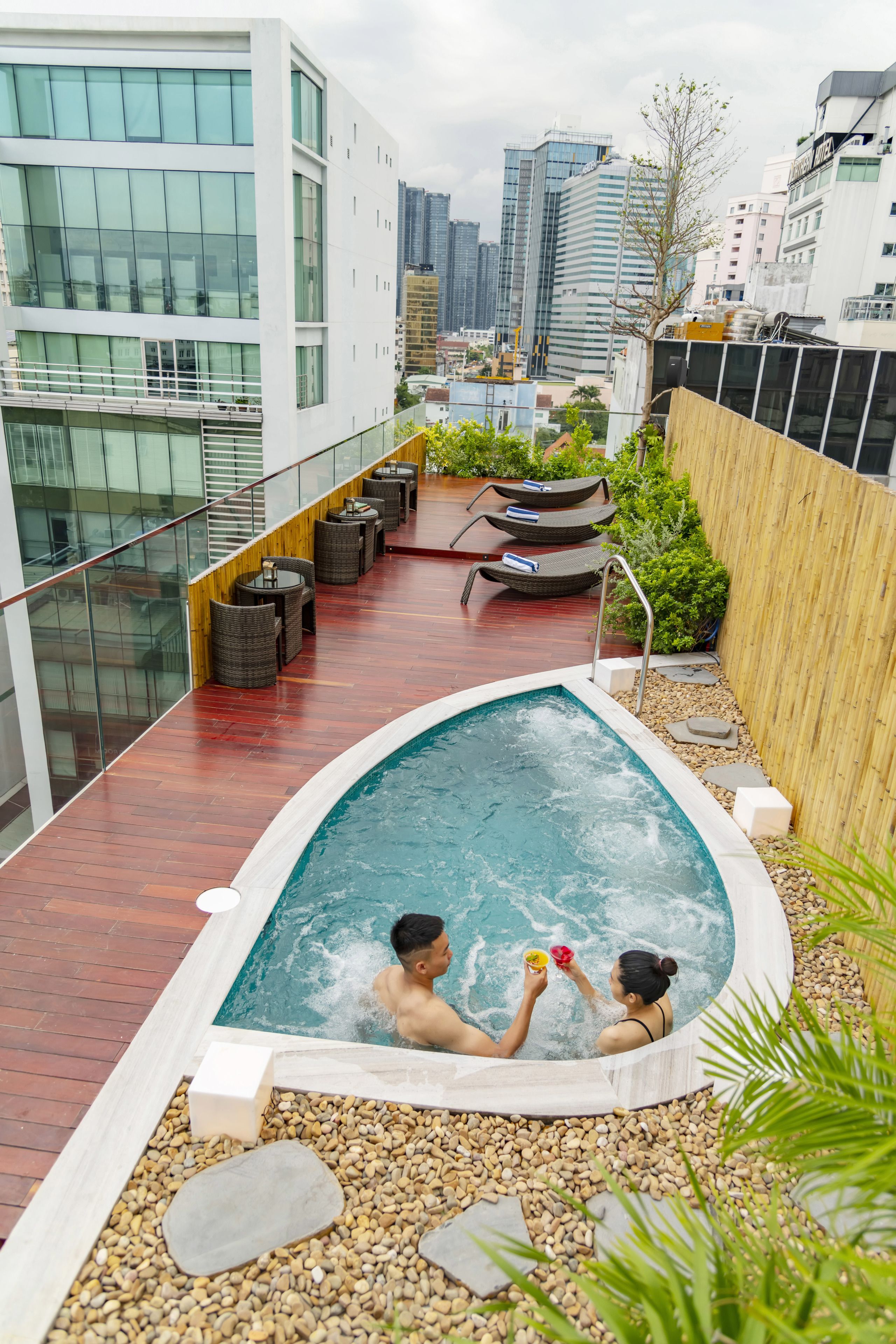 Piscina en la azotea con dos personas, rodeada de mobiliario exterior y vista panorámica de la ciudad.