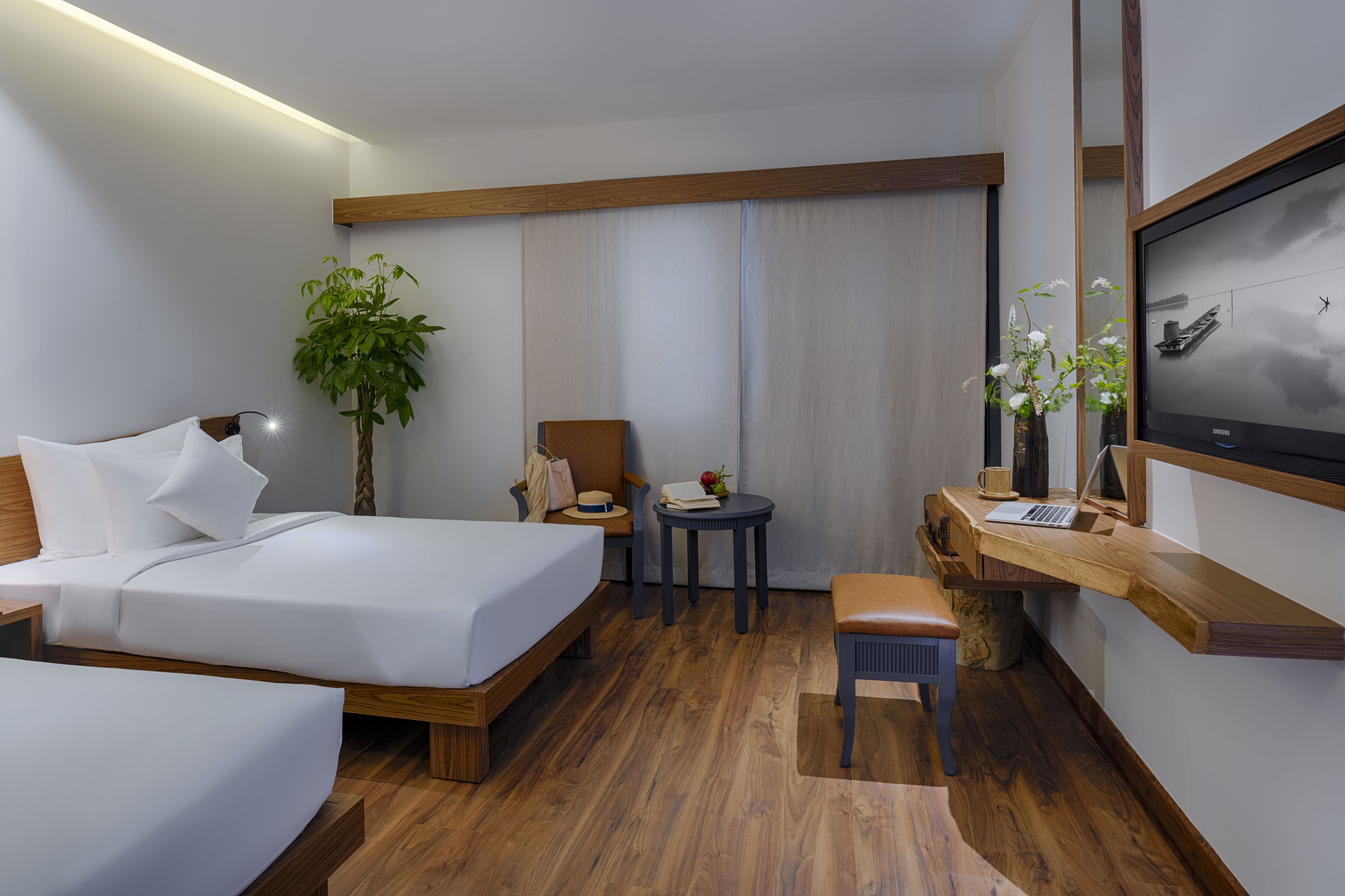 A hotel room with a bed, a desk with a laptop, a chair, a small table with a plant, and a television on the wall.