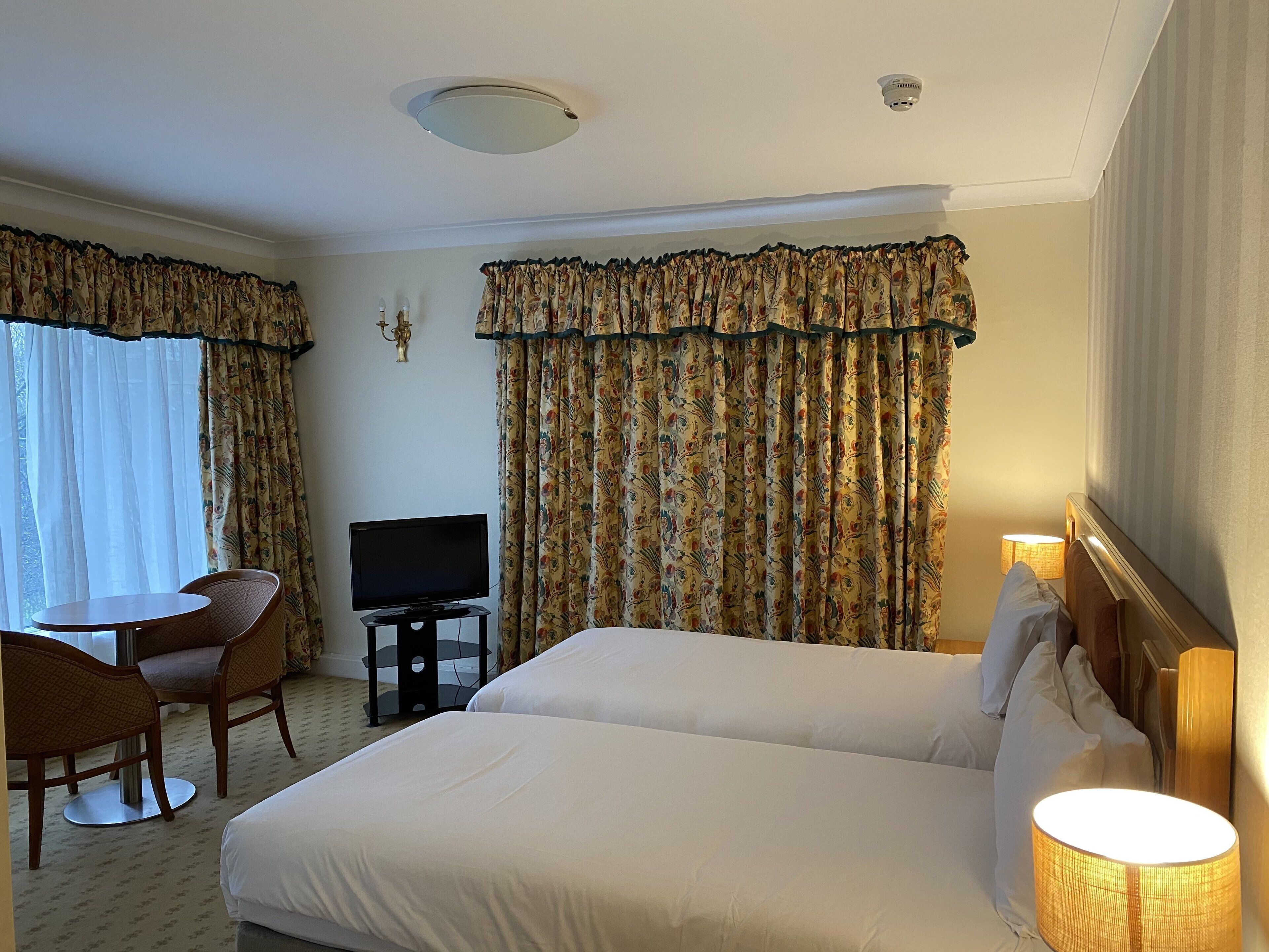 A hotel room with two beds, a TV, a small table with chairs, and curtains.