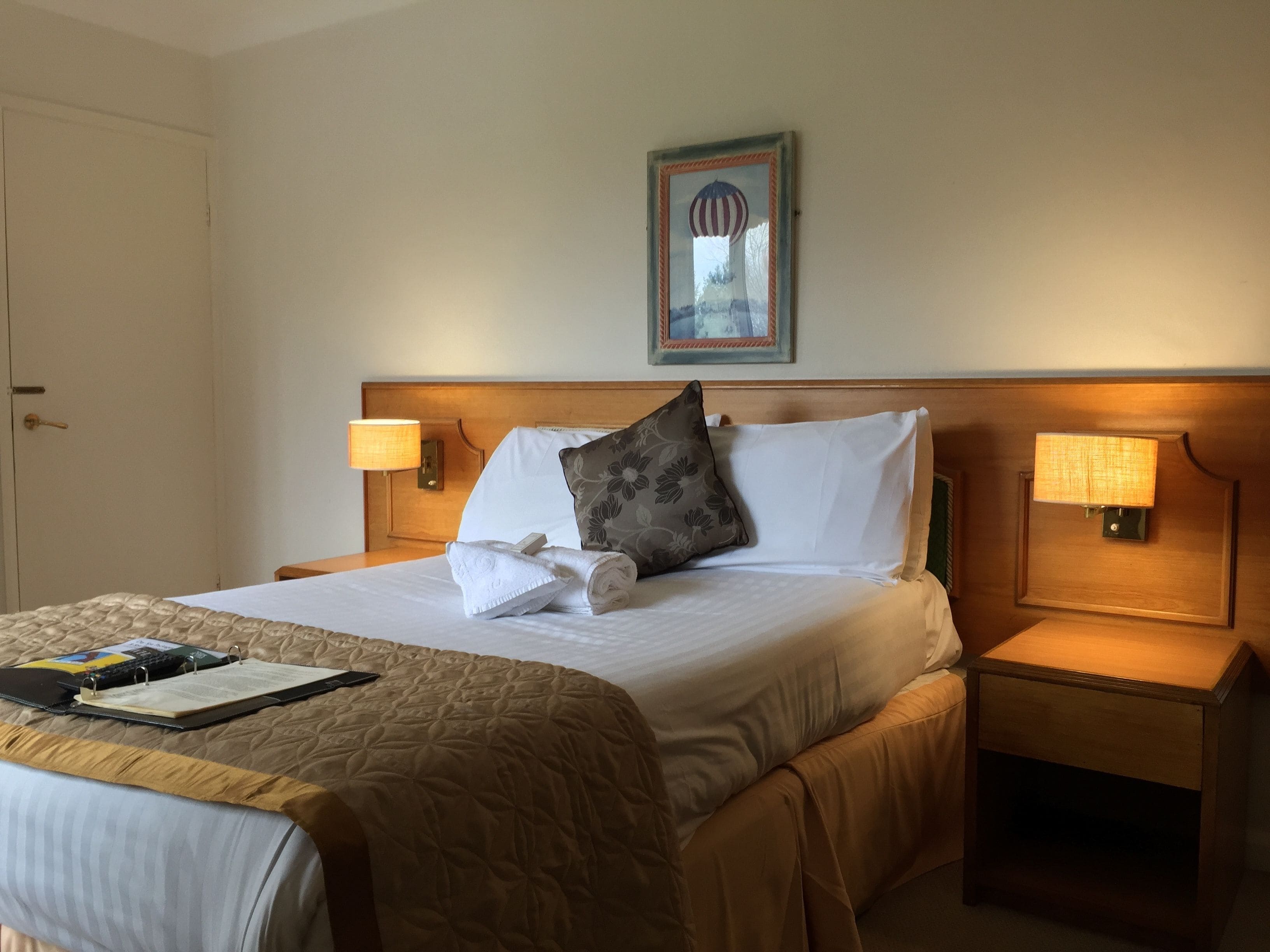 Habitación de hotel con una cama bien hecha, dos lámparas de noche, un cuadro en la pared y una puerta a la izquierda.