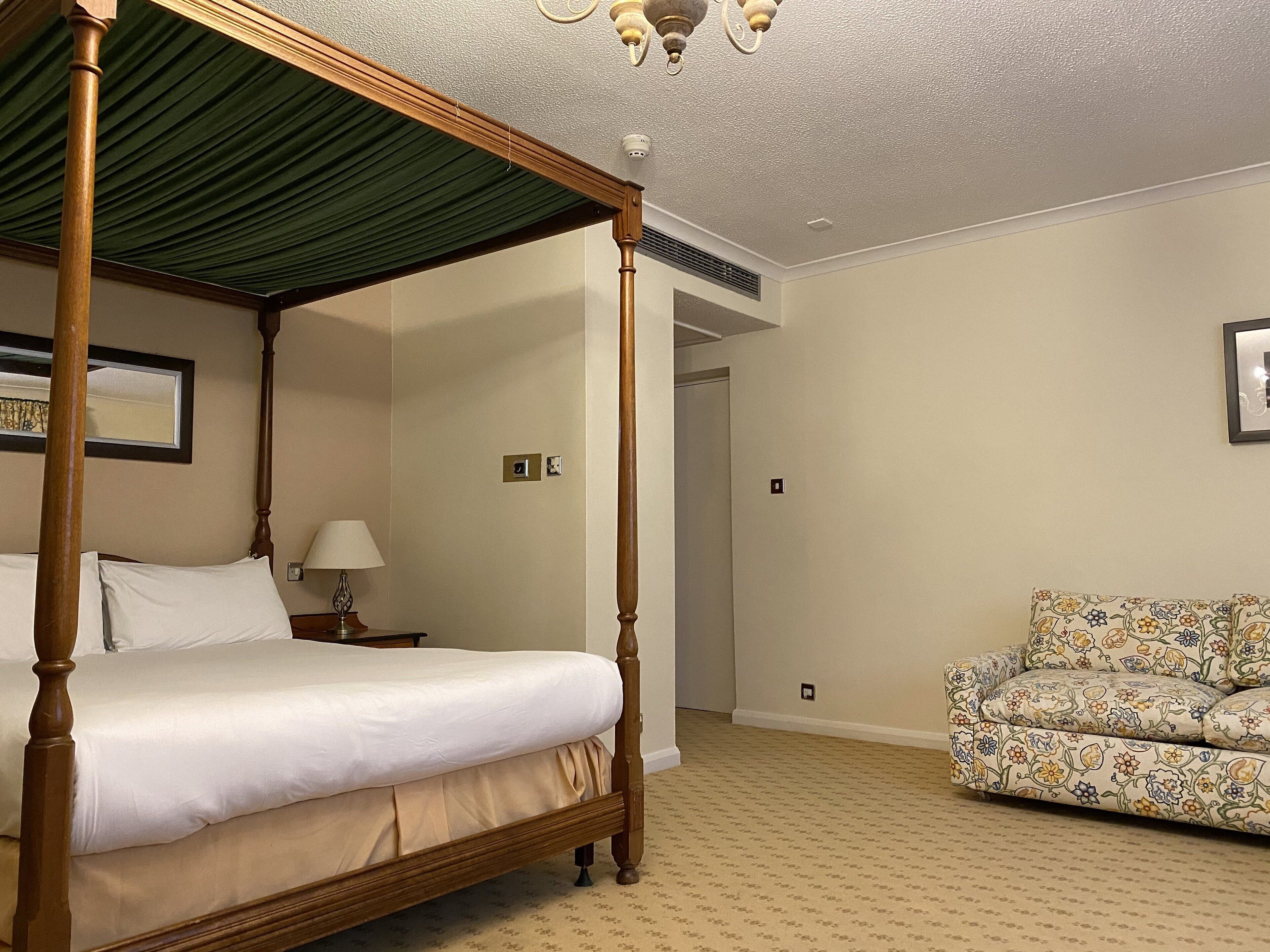 A four-poster bed with a green canopy, a bedside table with a lamp, a floral-patterned sofa, and a framed picture on the wall.
