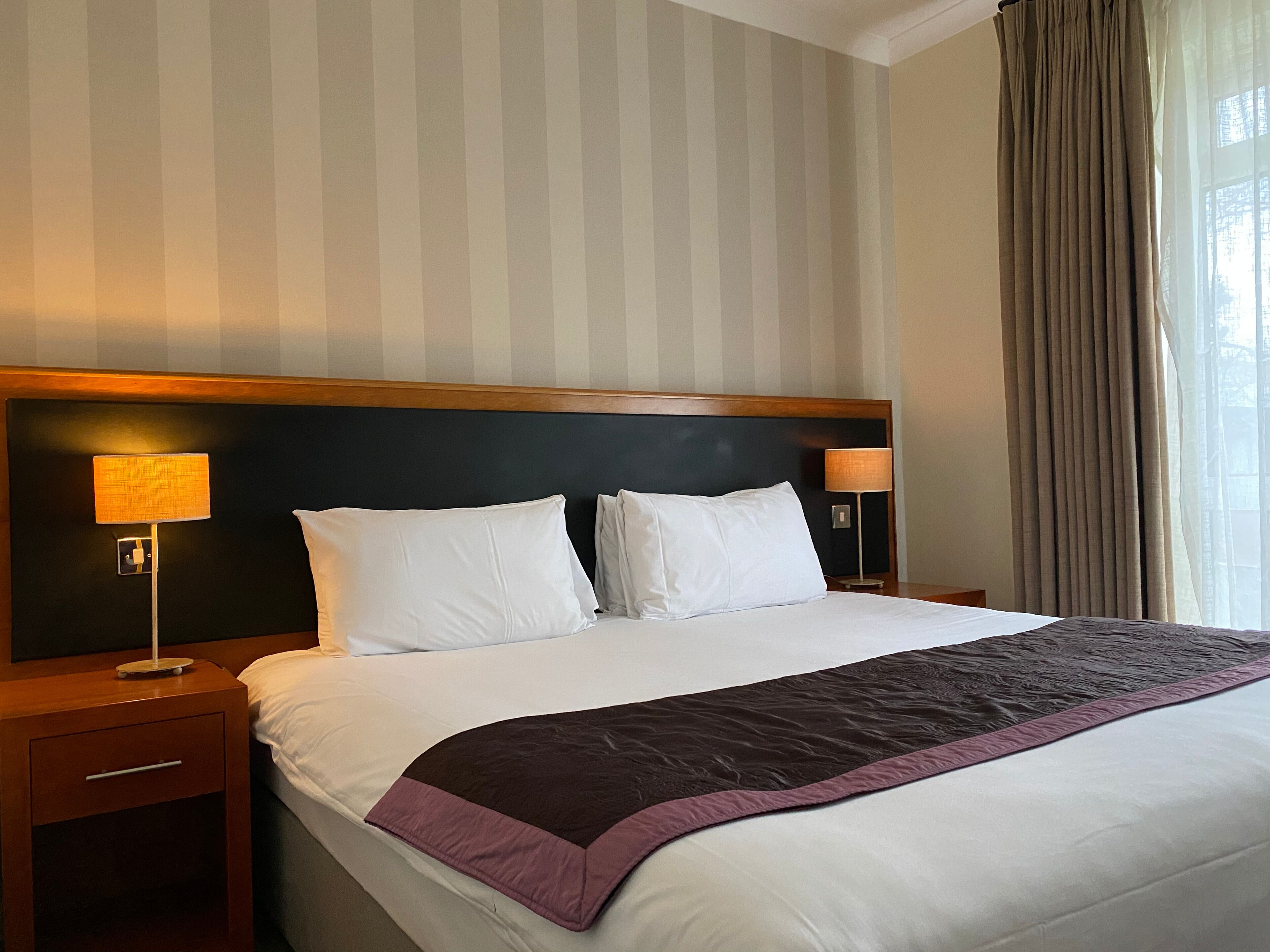 Habitación de hotel con una cama grande, dos lámparas de noche, un cabecero de madera y un ventanal con cortinas.