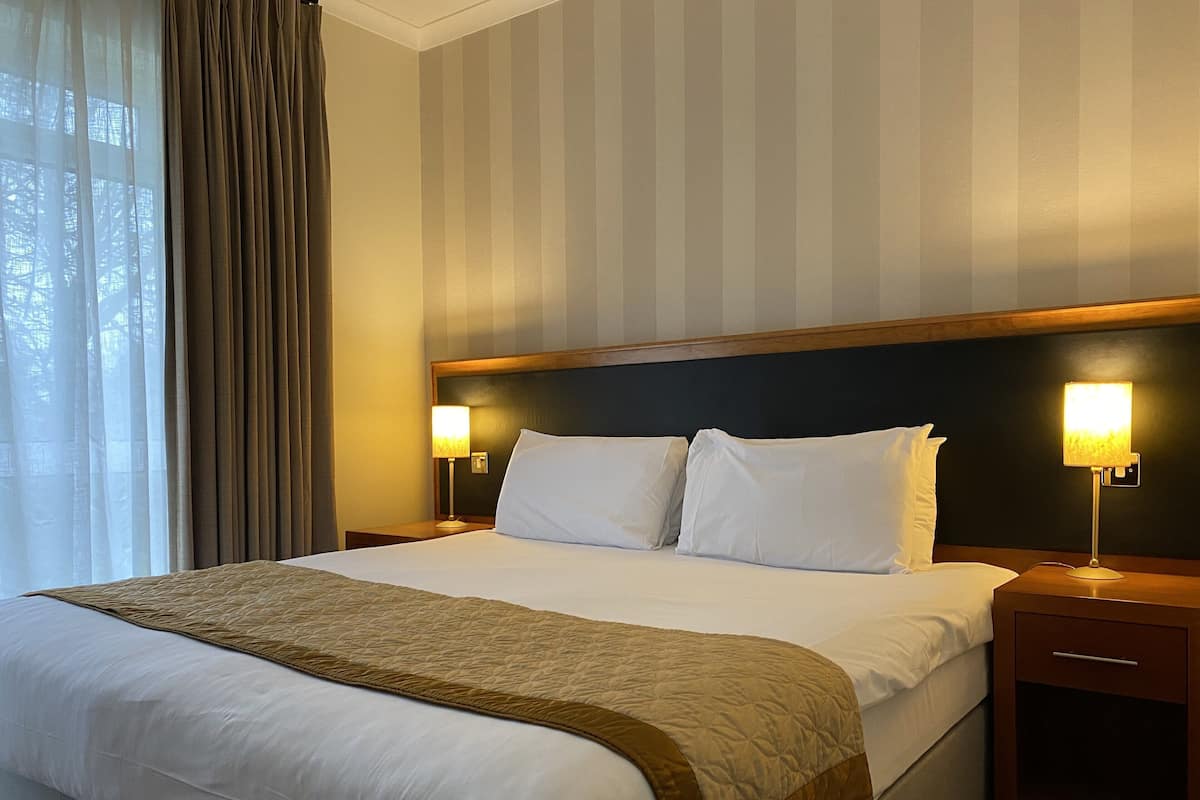 A hotel room with a large bed, two bedside tables, a headboard, and a window with curtains.
