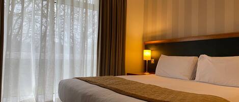 A hotel room with a large bed, a nightstand with a lamp, and sheer curtains on the window.