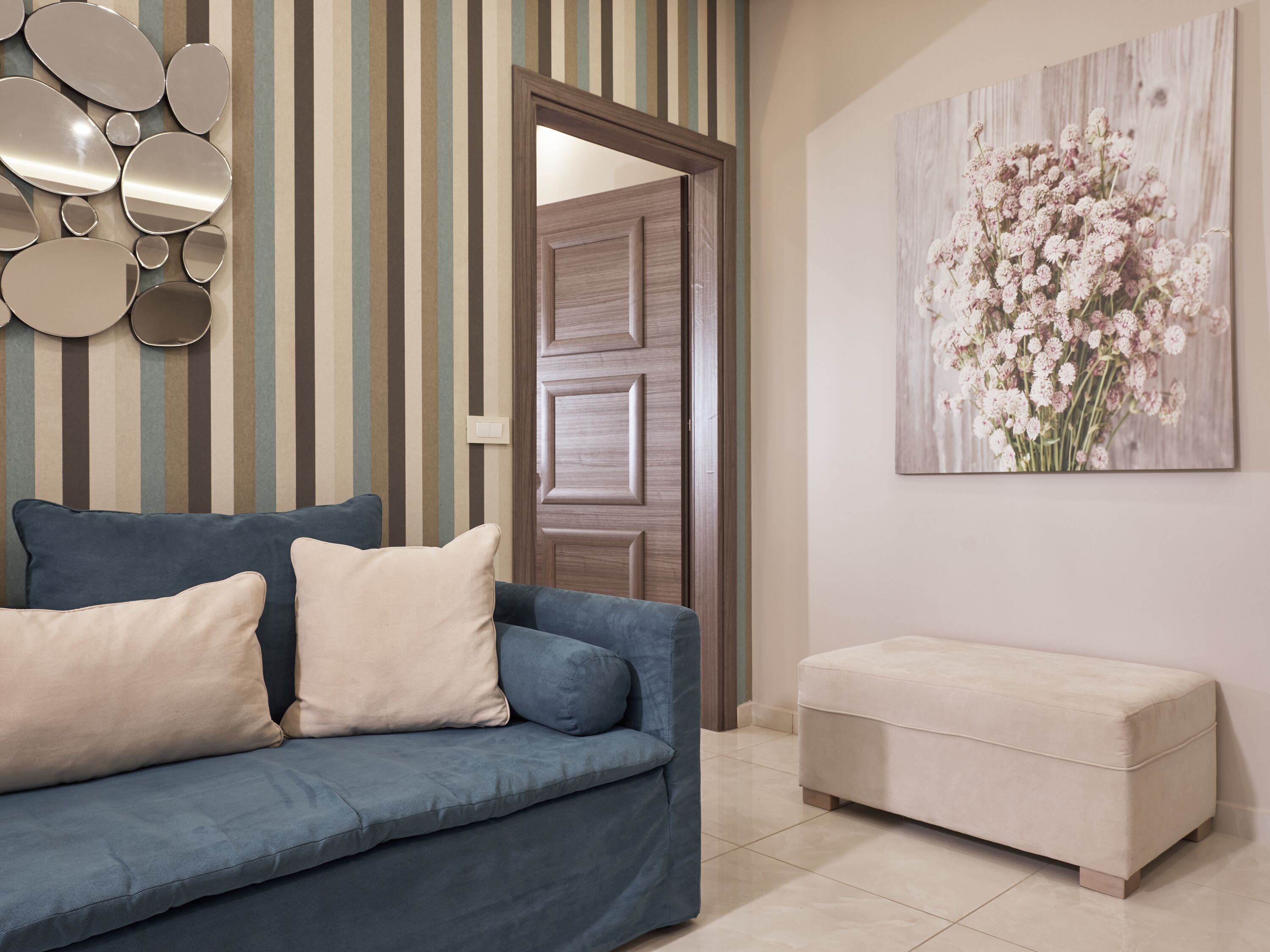 A modern living room with a blue sofa, beige cushions, a cream-colored ottoman, a wooden door, and a floral artwork on the wall.
