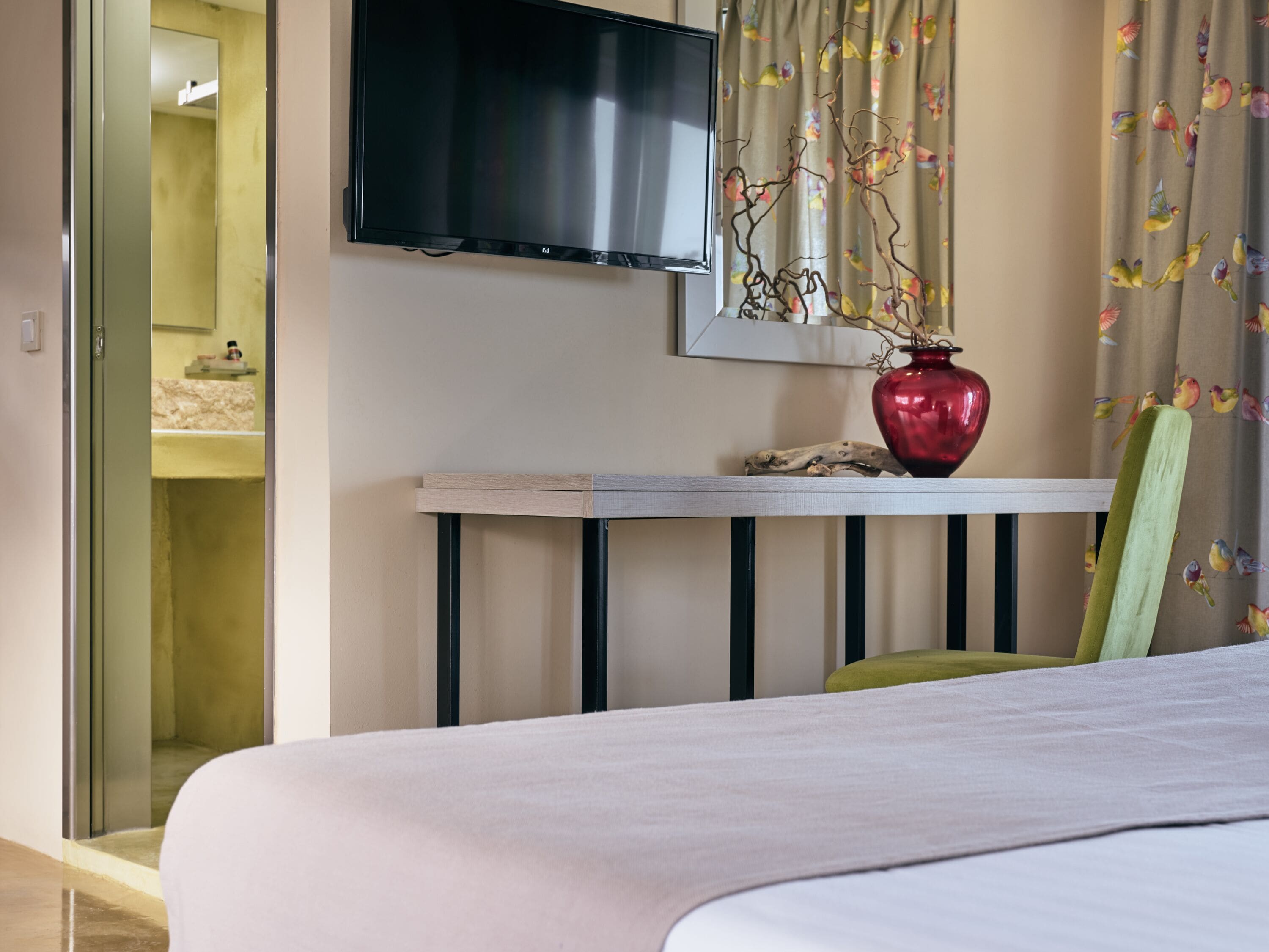 A hotel room with a bed, a TV mounted on the wall, a small table with a vase, and a curtain with a bird pattern.