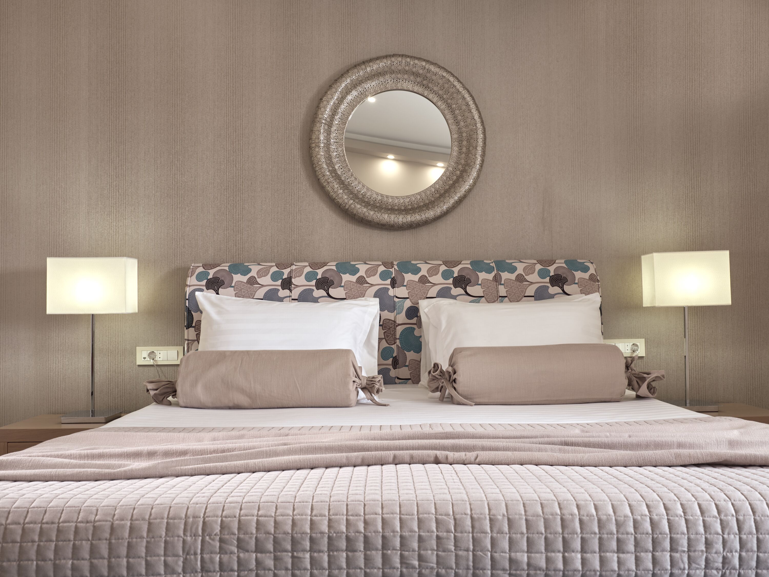 A neatly made bed with a quilted cover, two bedside lamps, and a decorative mirror on the wall.