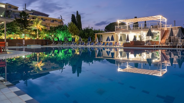 A swimming pool with lounge chairs and umbrellas, surrounded by buildings and trees. - Koukounaria Hotel & Suites (Zakynthos)