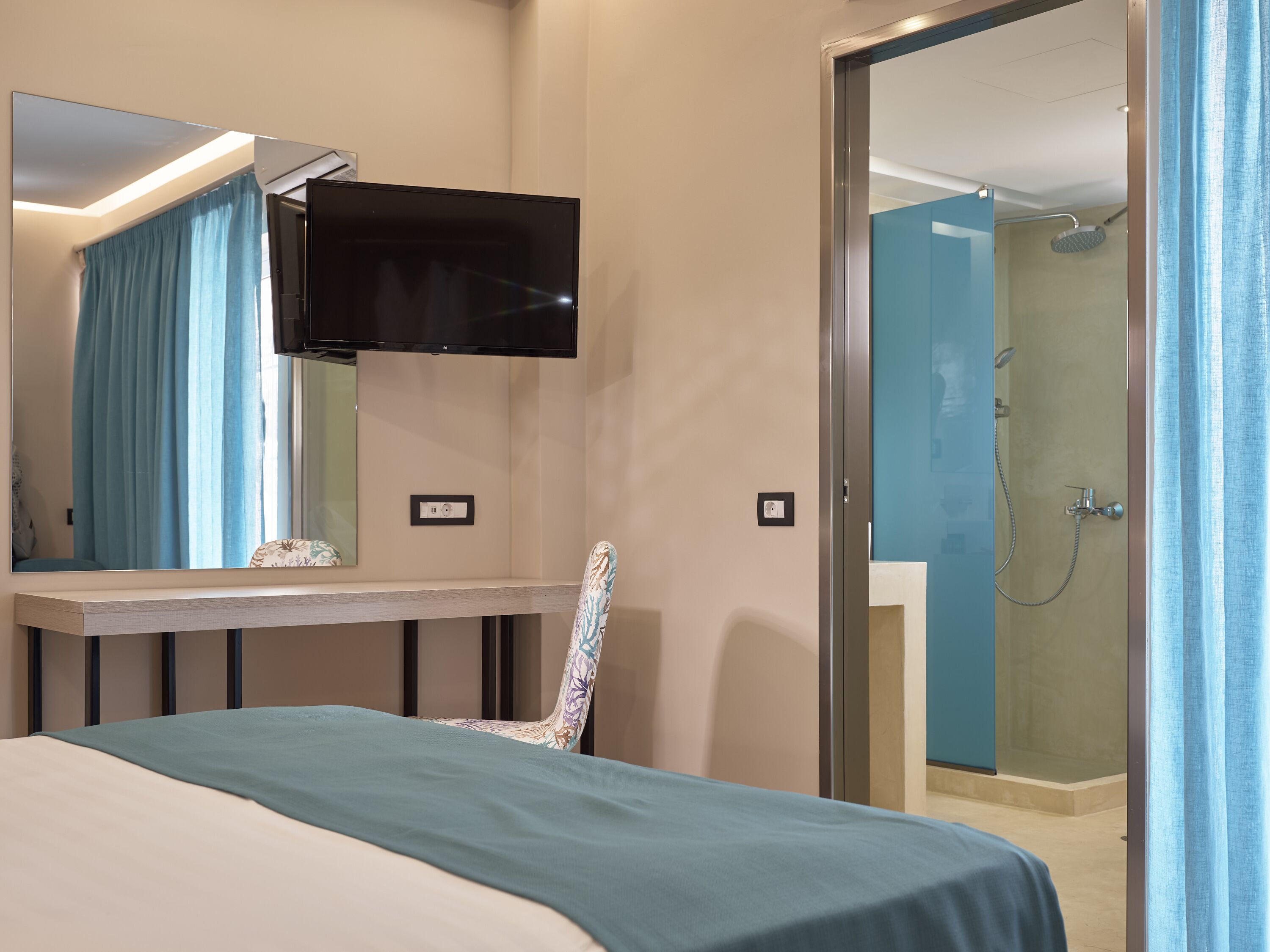 A hotel room with a bed, a desk with a flat-screen TV, a chair, a shower area, and a mirror.