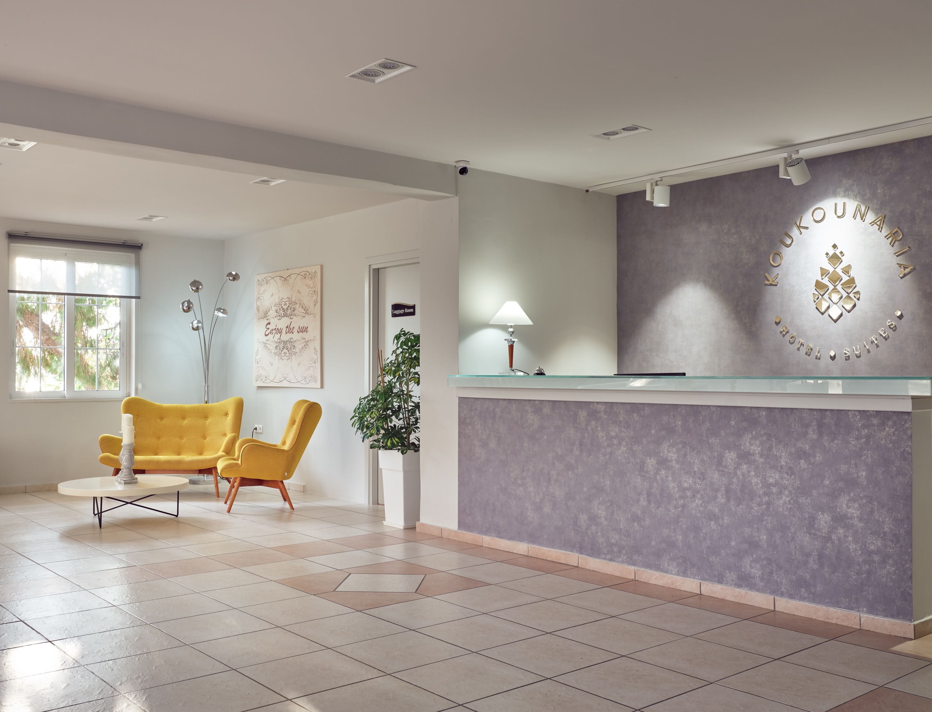 A hotel reception area with a modern design, featuring a reception desk, two yellow armchairs, a potted plant, and a sign indicating the lobby area.