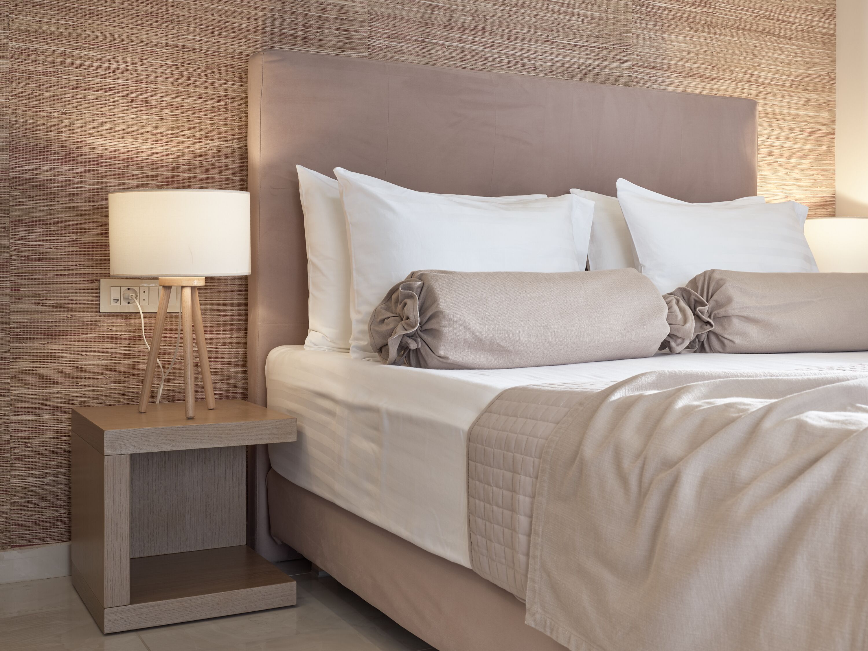 Un lit bien fait, avec une literie blanche et beige, une lampe de chevet et une petite table de nuit en bois.