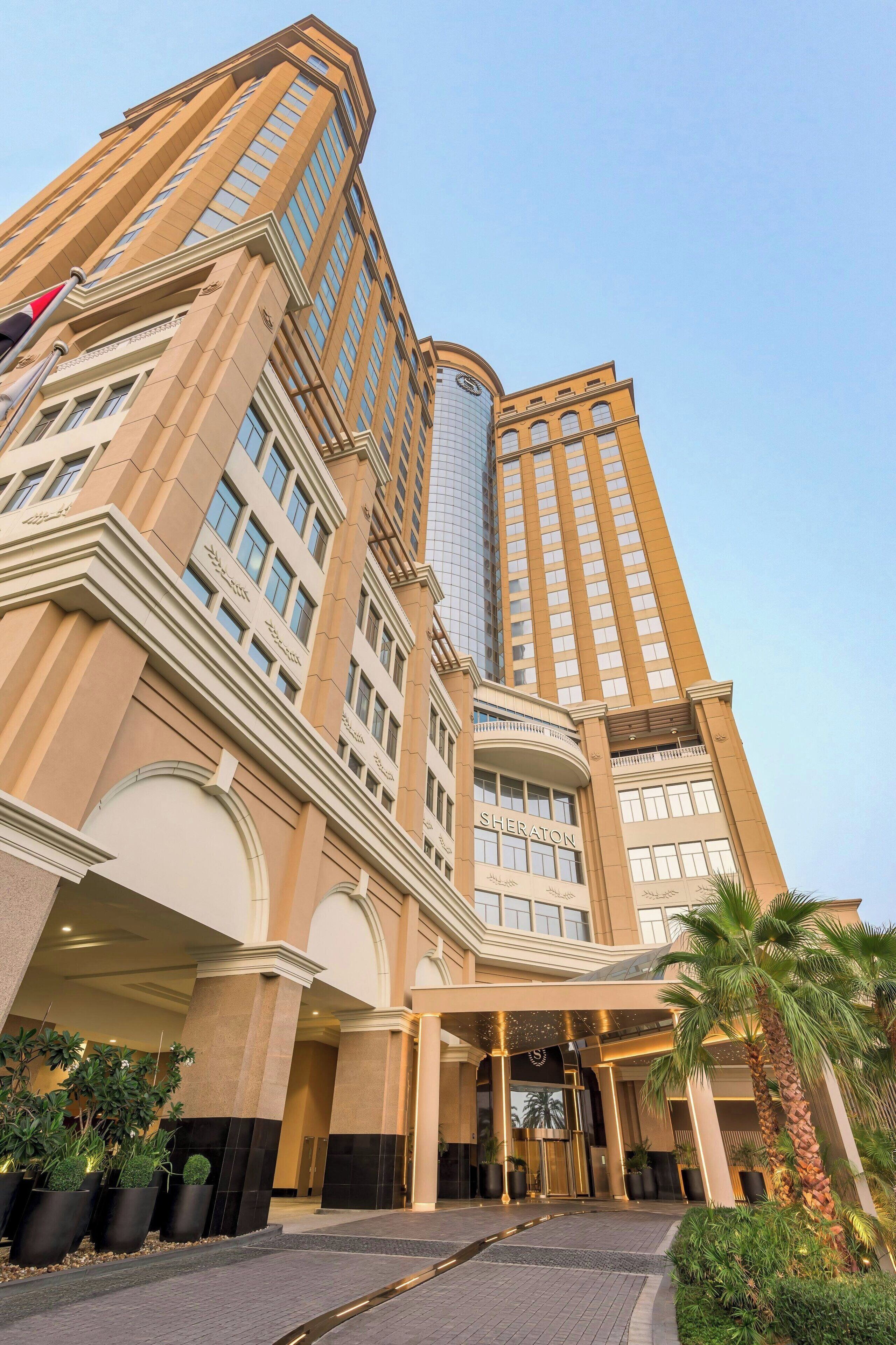 Un hotel de diseño moderno, con un llamativo letrero de Sheraton, un hueco de ascensor de vidrio y un área de entrada bien cuidada con palmeras y plantas en macetas.