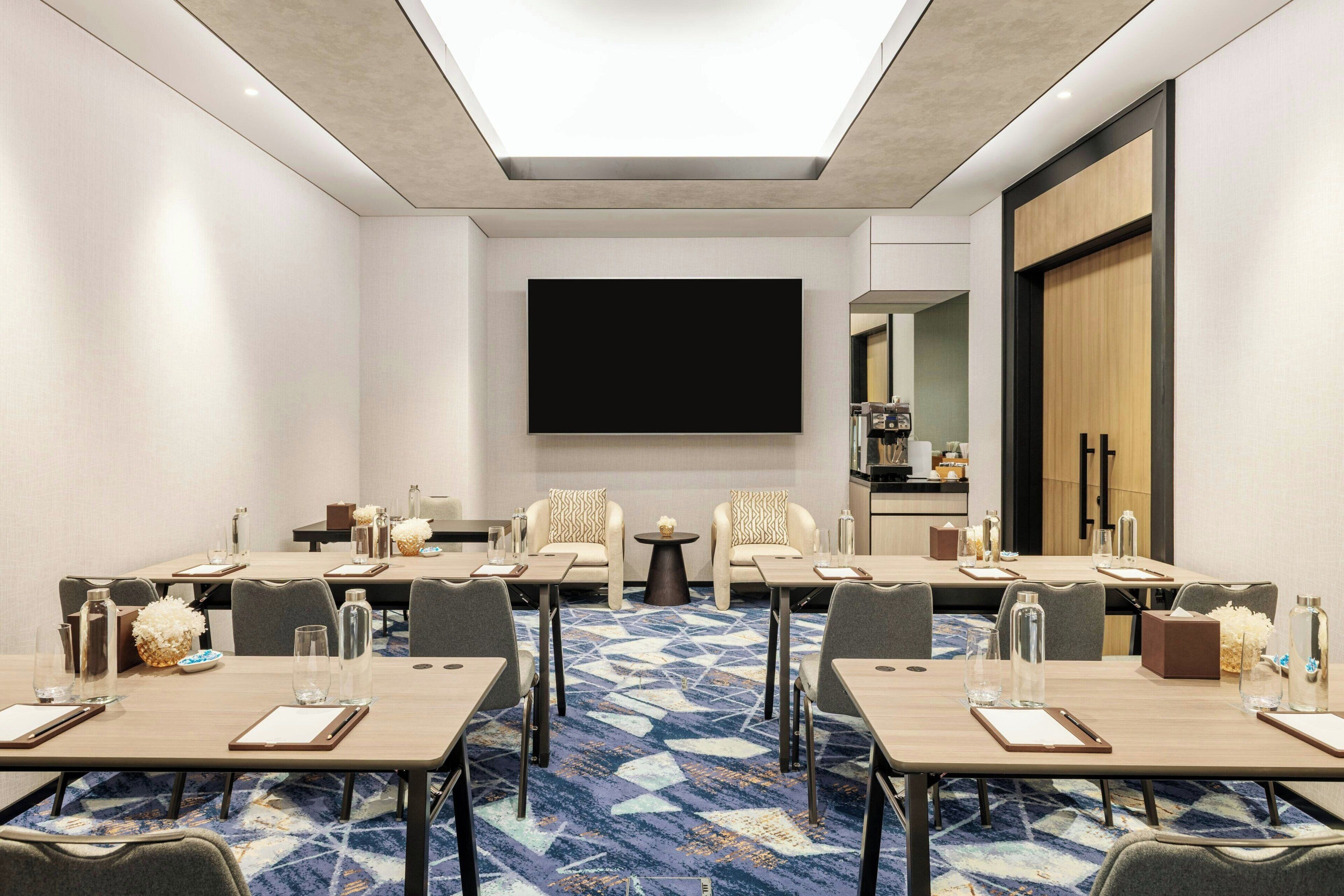 A conference room with a large flat-screen TV, multiple tables, and chairs, carpeted floor, and a modern ceiling design.