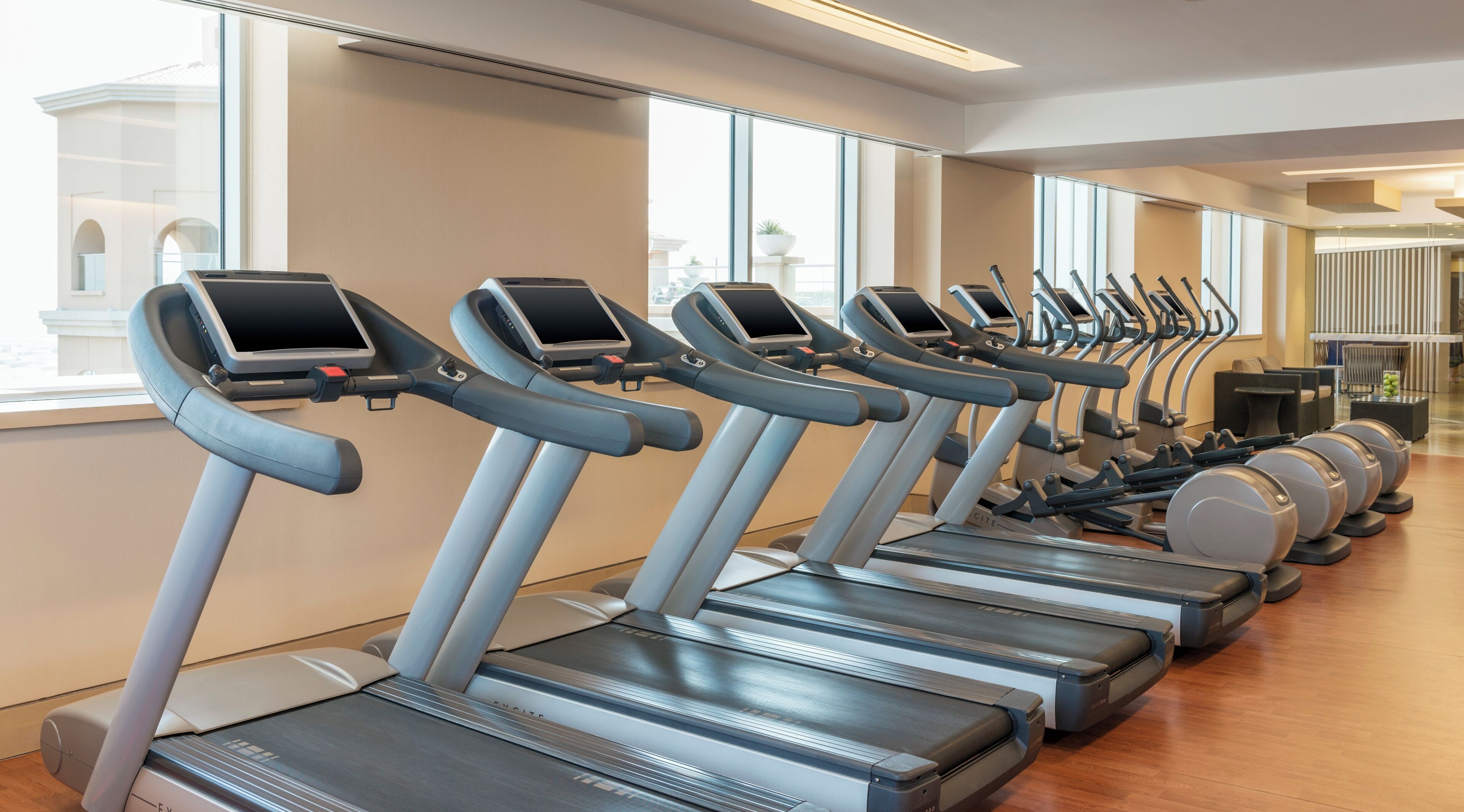 Una hilera de cintas de correr con pantallas digitales, suelo de madera y ventanas amplias.