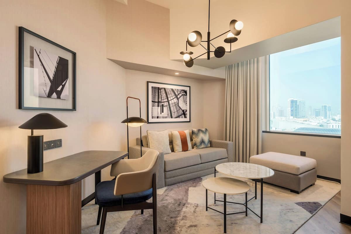Een moderne hotelkamer met een bank, een tafeltje, een bureau met lamp, en een groot raam met uitzicht over de stad.