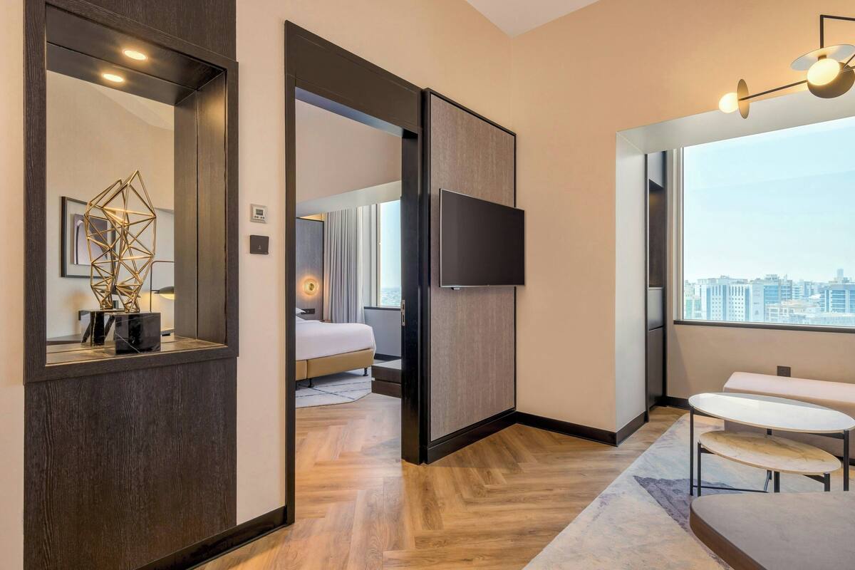 Een moderne hotelkamer met een grote spiegel, een flatscreen televisie, een houten vloer, een bed, een bankje en uitzicht op de stad.