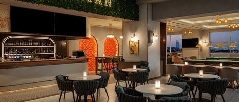 Zona de bar con un letrero que dice "SPARTAN", con barra surtida de botellas, mesas redondas con sillas e iluminación ambiental.