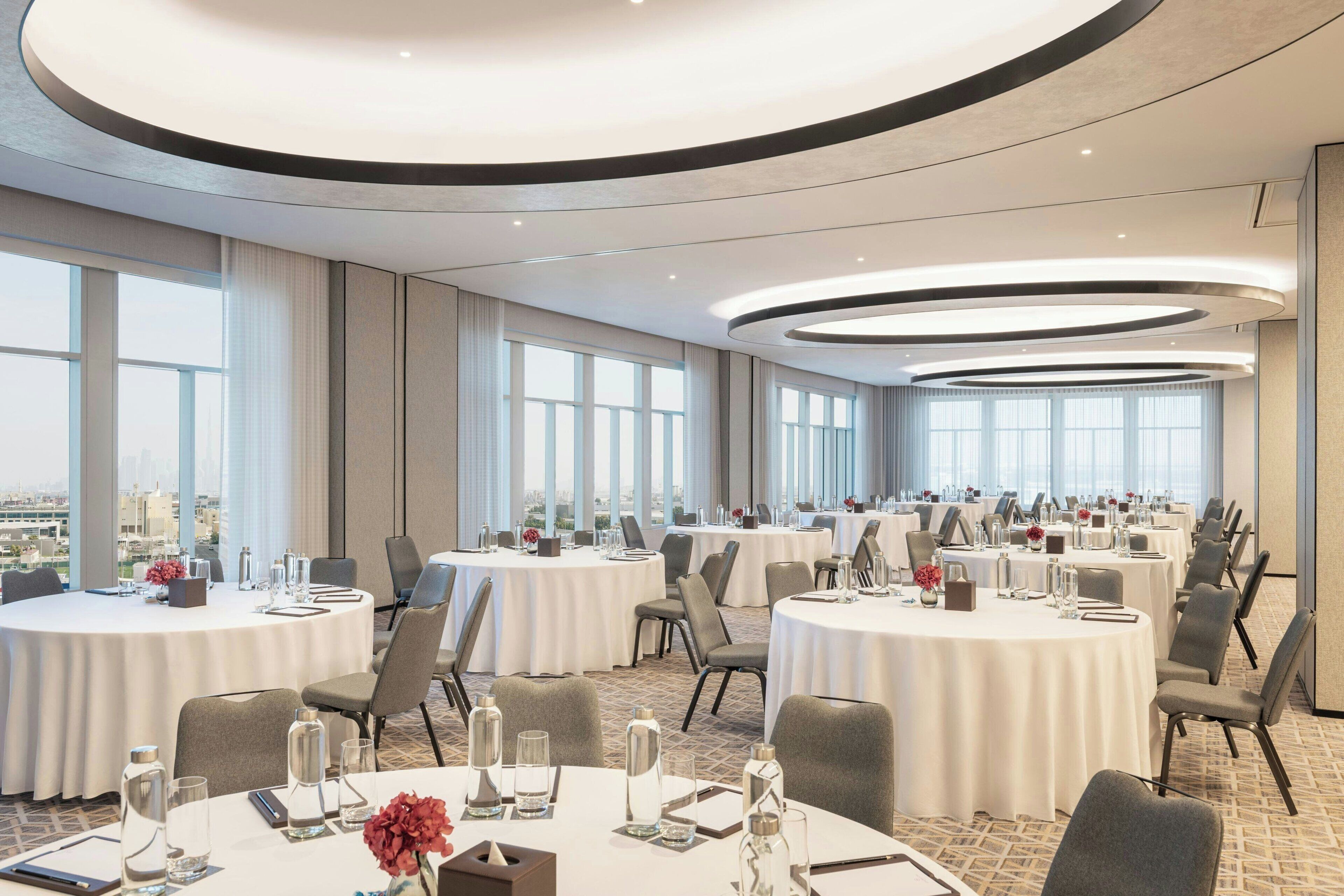Un salón de conferencias con varias mesas redondas, ventanas amplias y vista a la ciudad.
