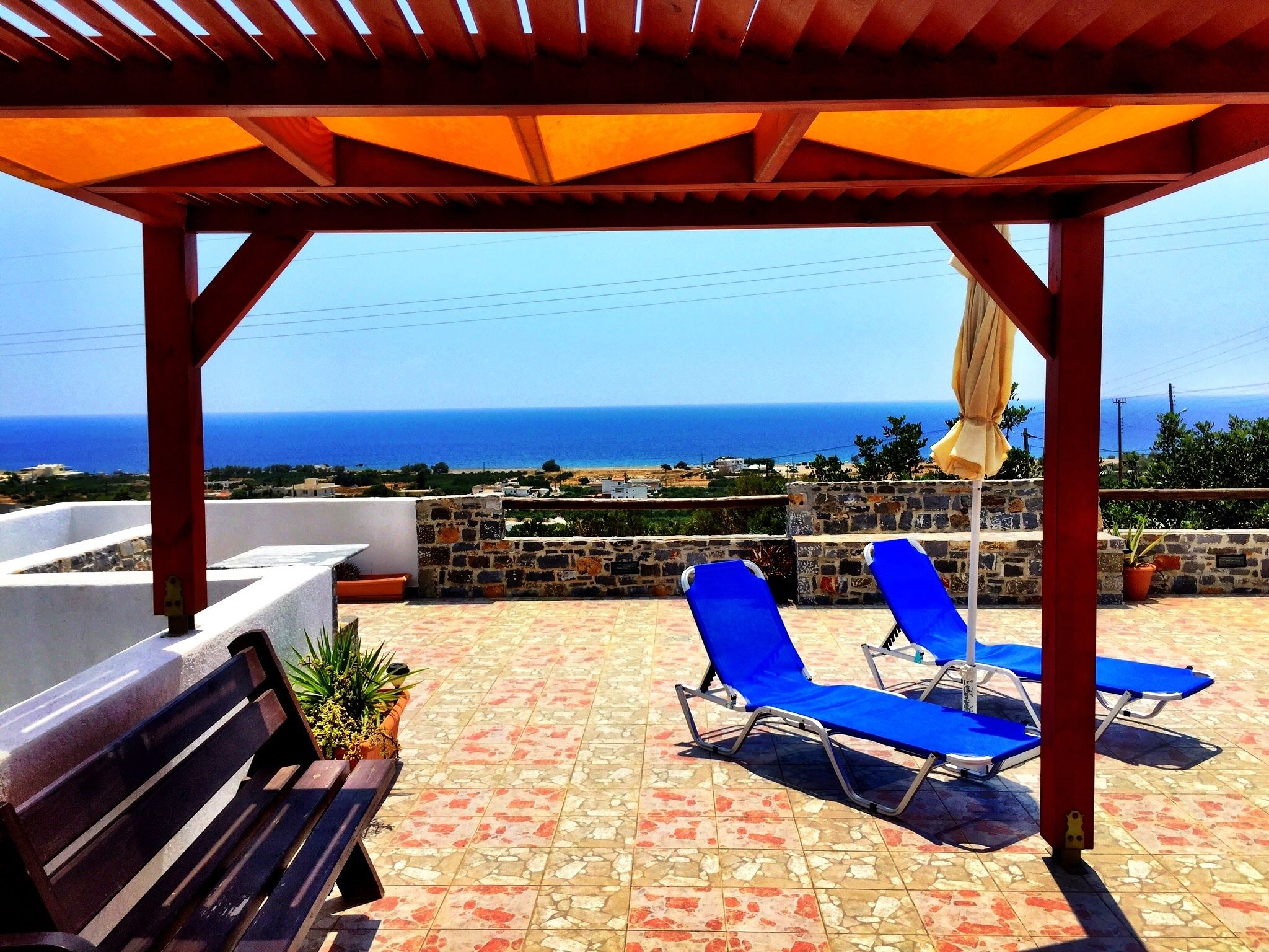A covered outdoor seating area with blue lounge chairs and a wooden bench, overlooking a coastal view.