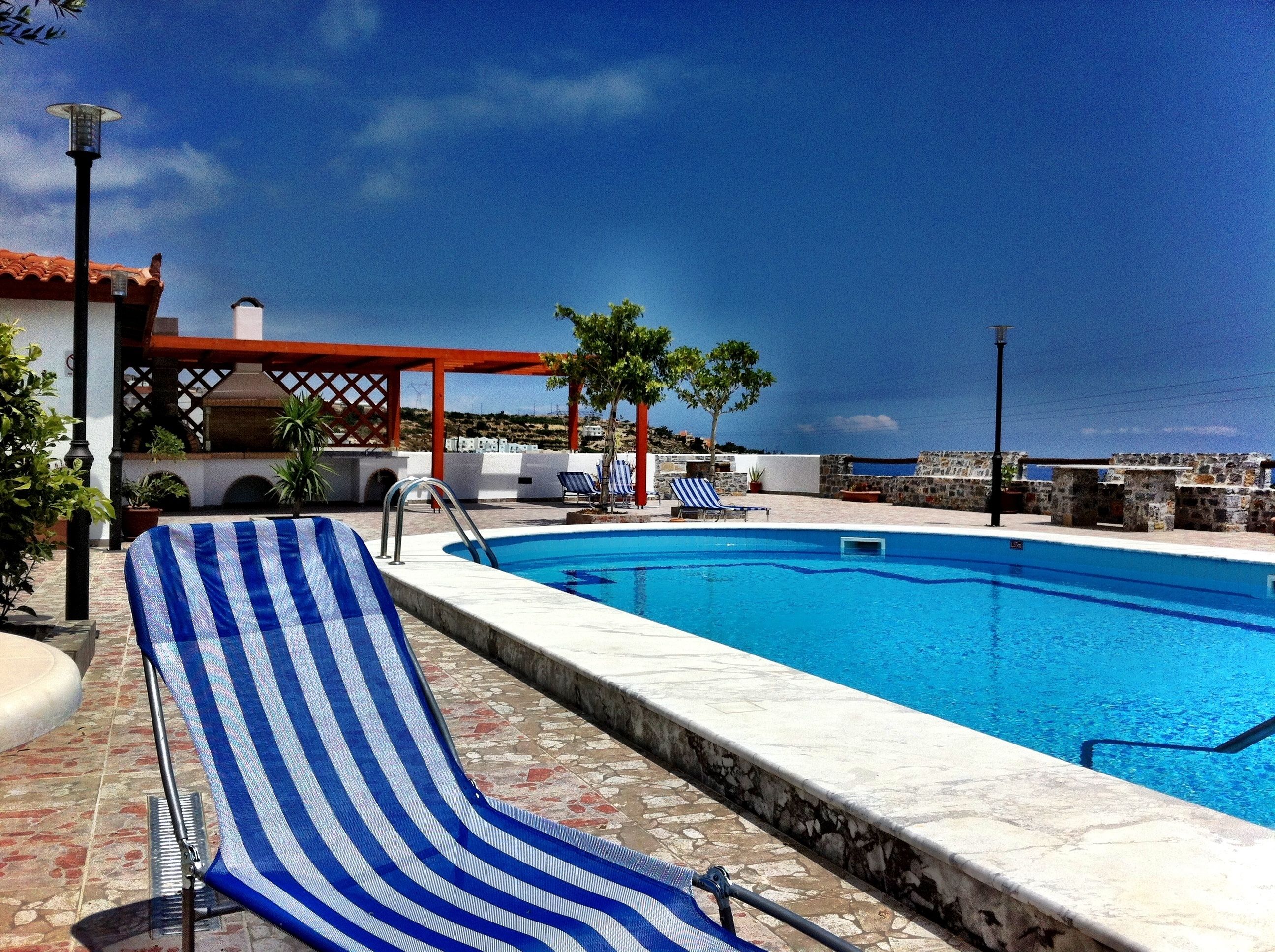 A pool area with a clear blue pool, lounge chairs, and a view of the sea.