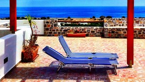 Una pérgola roja con dos sillones a rayas y una planta en maceta en un patio empedrado con vistas al mar.