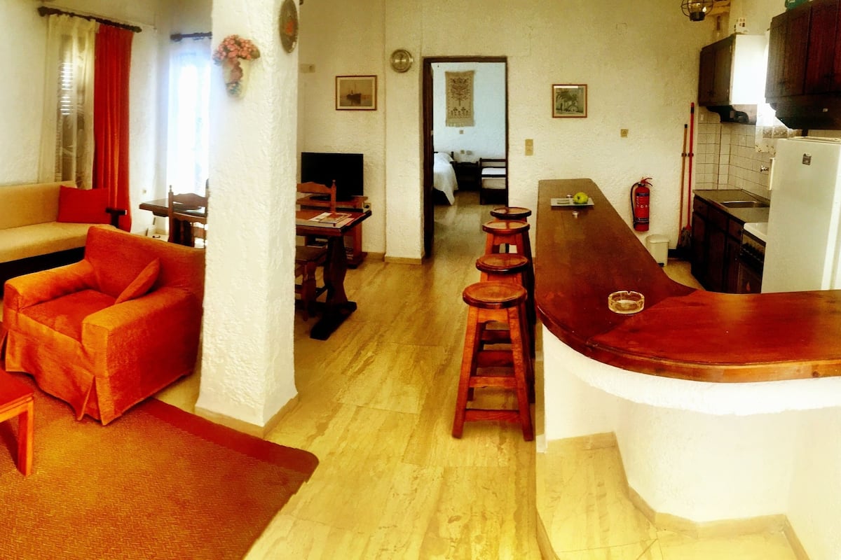 A living room with a sofa, chairs, a coffee table, and a dining area with a table and chairs. There is a kitchen area with a refrigerator, sink, and cabinets.