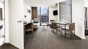 A hotel room with a dining area, a sofa, a desk, a TV, and a view of the city.