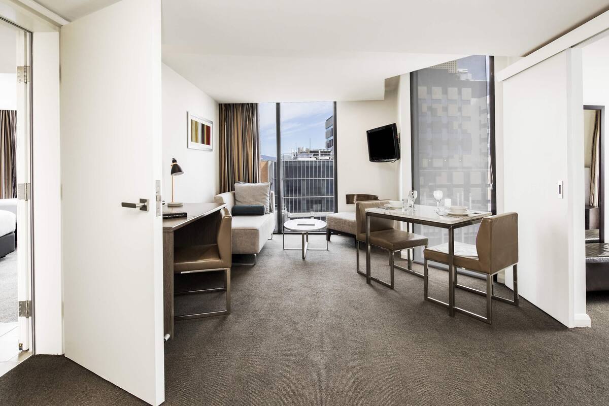 Quarto de hotel com área de refeições, sofá, escrivaninha, televisão e vista para a cidade.