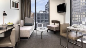 A modern hotel room with a sofa, armchair, dining table, and a TV on the wall.