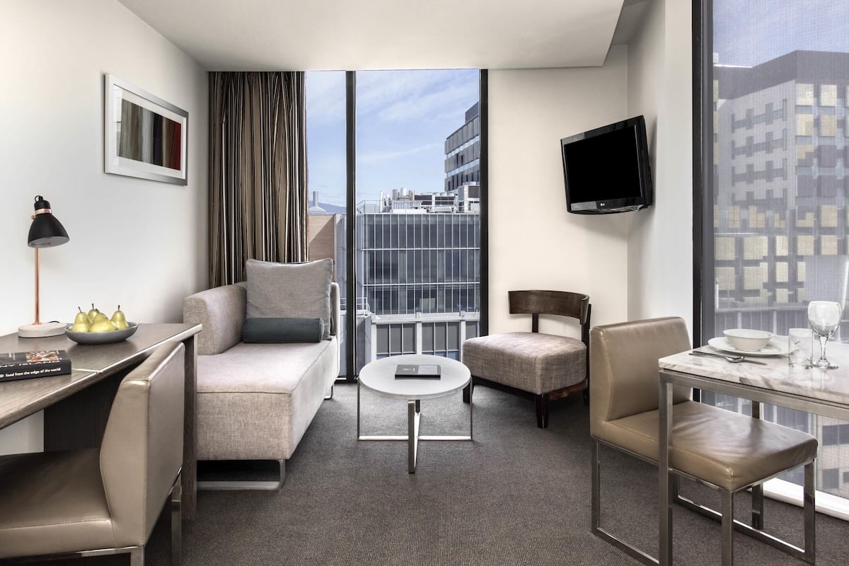 Quarto de hotel moderno com sofá, poltrona, mesa de jantar e televisão na parede.