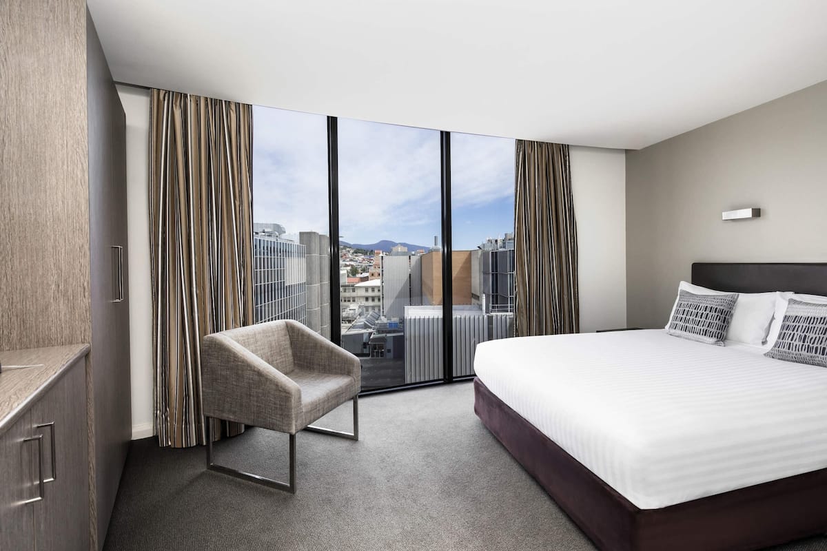 Quarto de hotel com uma cama grande, uma cadeira, um guarda-roupa e vista da cidade através de janelas do chão ao teto.