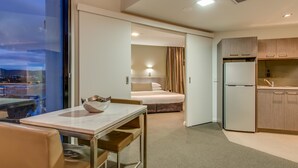 A modern hotel room with a dining area, a bed, a kitchen, and a view of the cityscape.