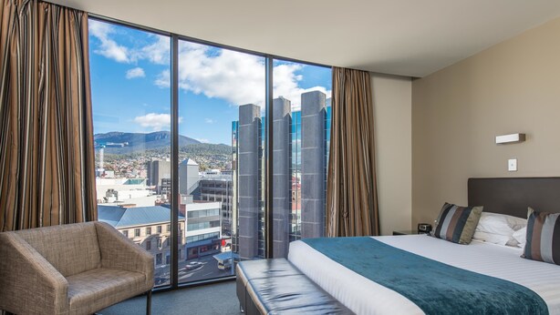 A hotel room with a large window, a bed, a chair, and a bench.