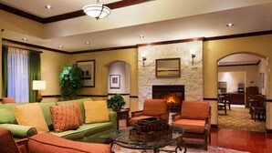 A spacious living room with a green sofa, orange armchairs, and a glass coffee table. A fireplace is visible in the background, and there is a dining area with wooden furniture. - Country Inn & Suites by Radisson, College Station, TX (College Station)