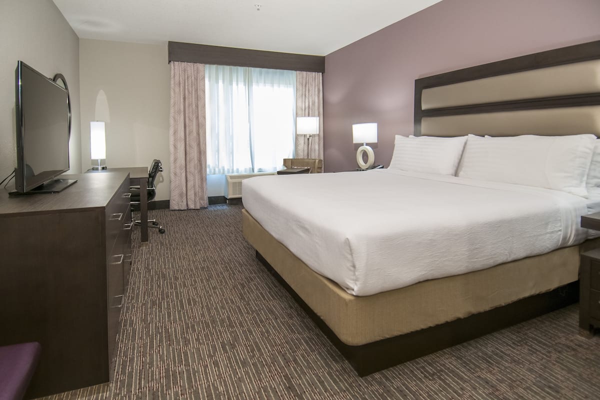 Habitación de hotel con una cama grande, un escritorio con televisor de pantalla plana, una silla, dos lámparas de noche y una ventana con cortinas.