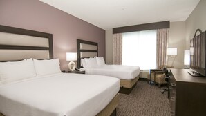 Habitación de hotel con dos camas, un escritorio, una silla, una lámpara y un ventanal con cortinas.