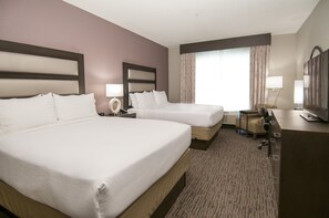 Habitación de hotel con dos camas, un escritorio, una silla, una lámpara y un ventanal con cortinas.