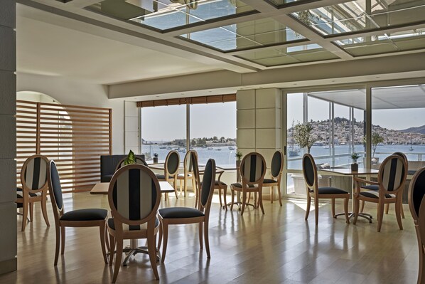 A spacious room with wooden chairs and tables, large windows offering a view of a body of water and a distant hill. - Xenia Poros Image Hotel (Poros)