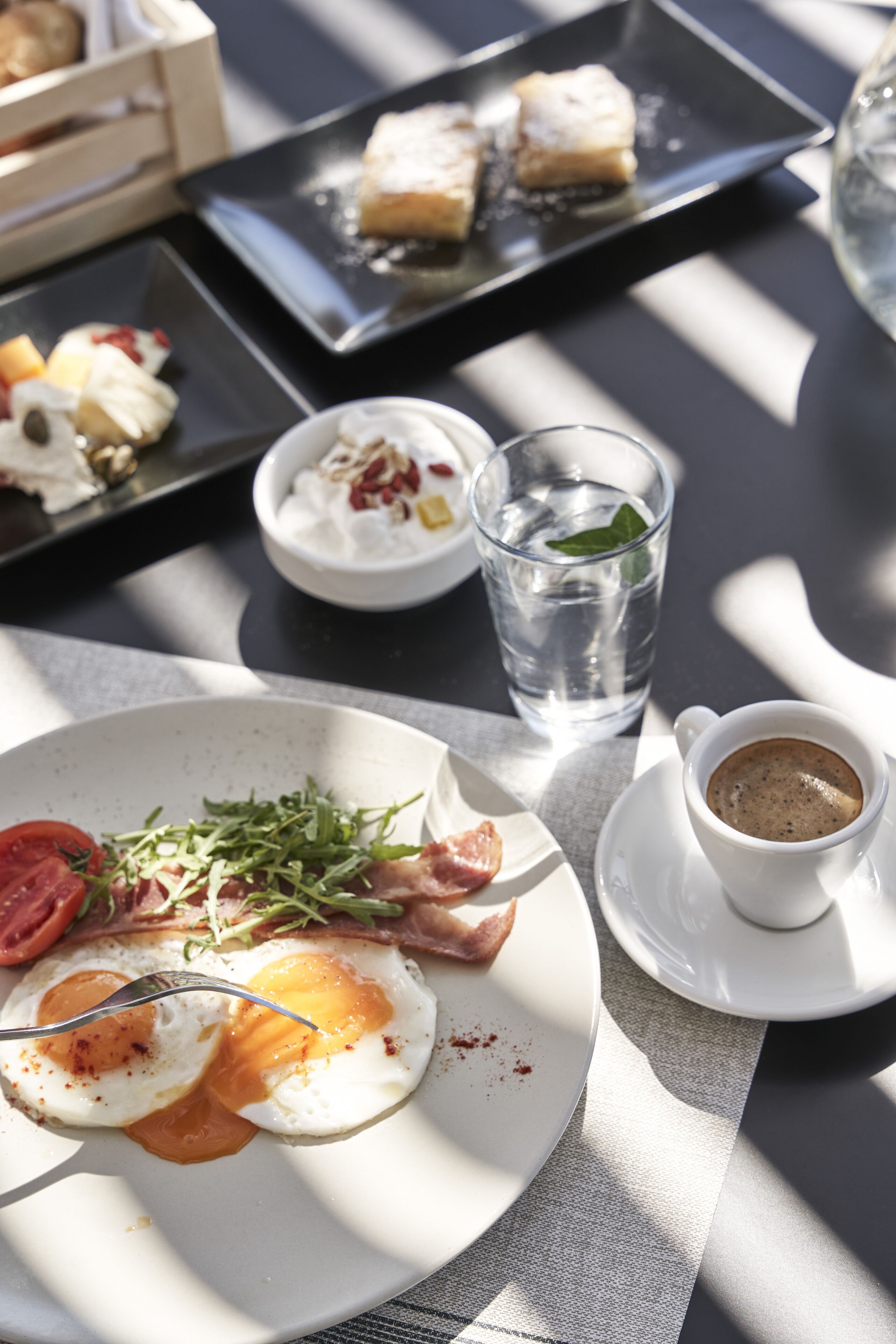 A breakfast setting with a plate of eggs, bacon, and greens, a glass of water, and a cup of coffee on a table.