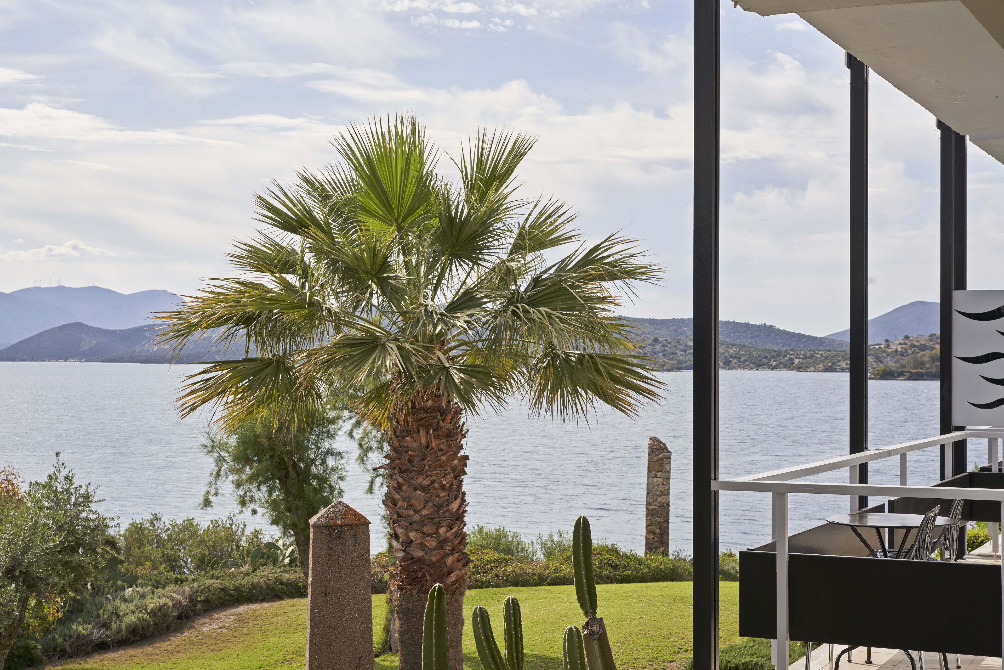 A view from a balcony overlooking a lake with mountains in the background, a palm tree, and a patio area with a table and chairs.