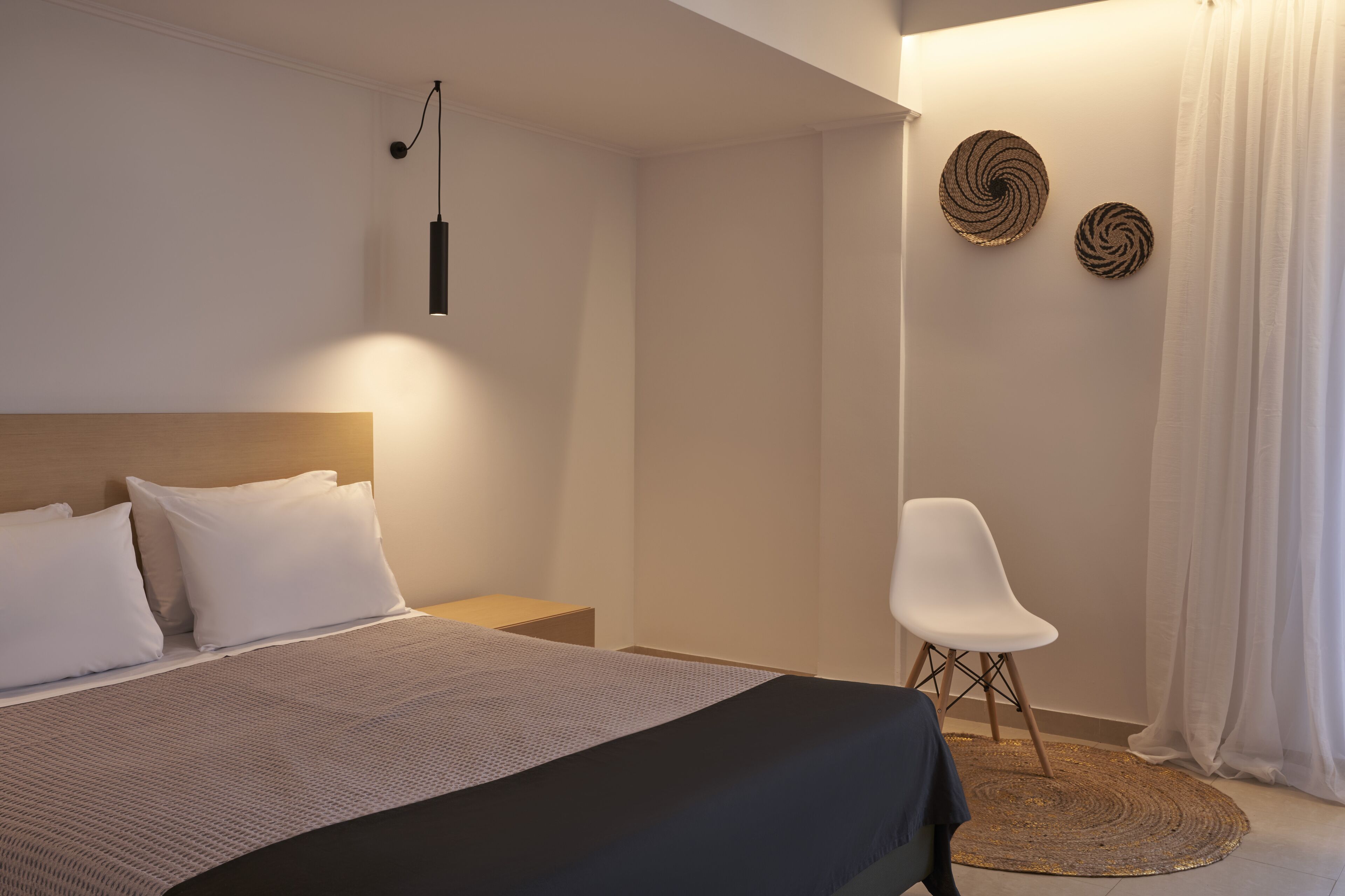 A modern hotel room with a neatly made bed, a bedside table, a chair, and two wall-mounted decorative items.