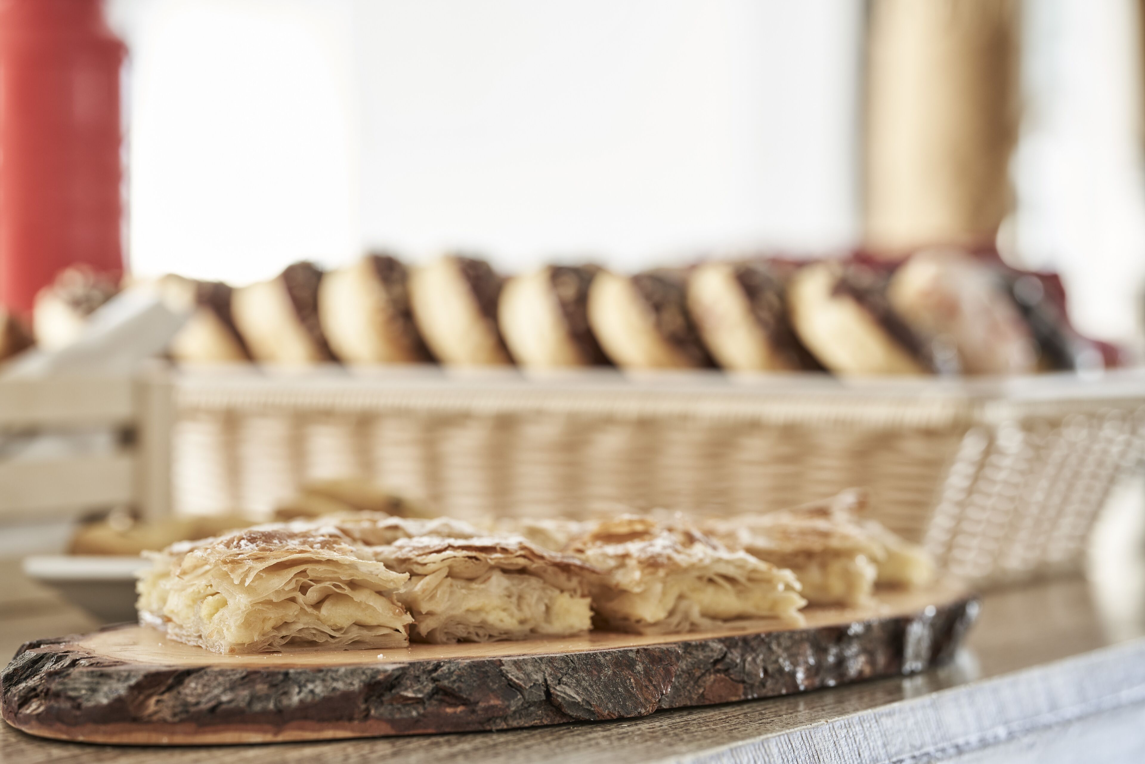 A wooden cutting board with pastry on it, a wicker basket of pastries in the background, and a red object to the left.