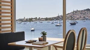 A view from a window overlooking a harbor with boats, a cityscape, and a table with a plant, book, and drink. - Xenia Poros Image Hotel (Poros)