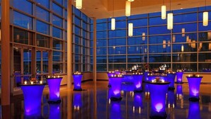 A spacious indoor area with multiple tables illuminated by blue lighting, large windows, and hanging light fixtures.
