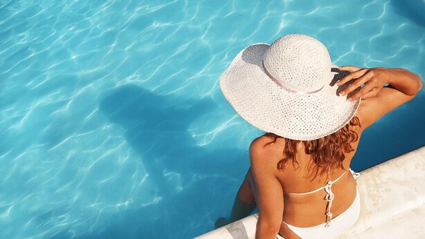 A person sitting by a pool with clear blue water, wearing a white bikini and a large straw hat.