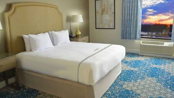 A hotel room with a large bed, two bedside tables with lamps, a window with a view of a sunset, and a patterned carpet.