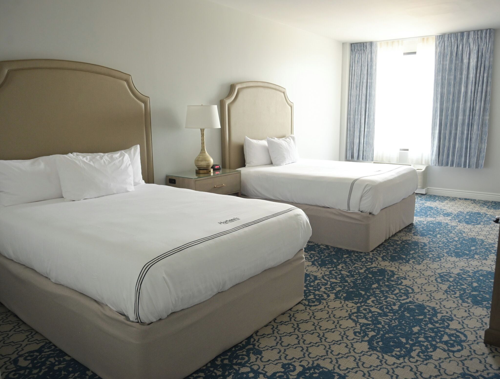 A hotel room with two beds, a nightstand with a lamp, a window with curtains, and a patterned carpet.